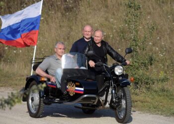 Vladimir Putin montando una motocicleta con un sidecar, acompañado por dos hombres y una bandera rusa.