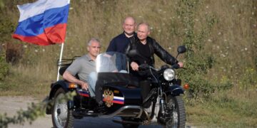 Vladimir Putin montando una motocicleta con un sidecar, acompañado por dos hombres y una bandera rusa.