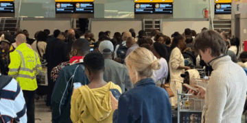 Una terminal de aeropuerto llena de gente con pasajeros en la fila y pantallas que muestran "Escritorio cerrado."