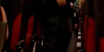 Un hombre con un uniforme oscuro escolta a un pasajero agarrando una botella de vodka por un pasillo de avión.