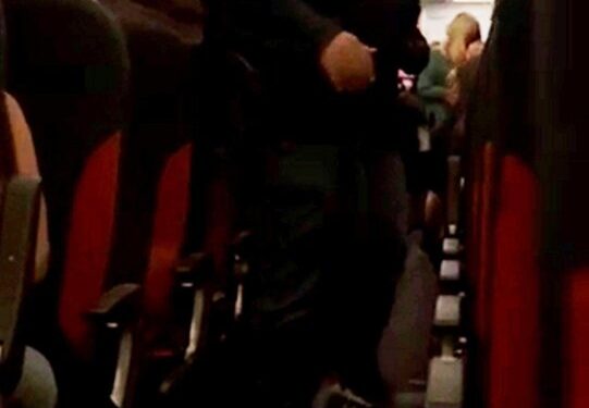 Un hombre con un uniforme oscuro escolta a un pasajero agarrando una botella de vodka por un pasillo de avión.