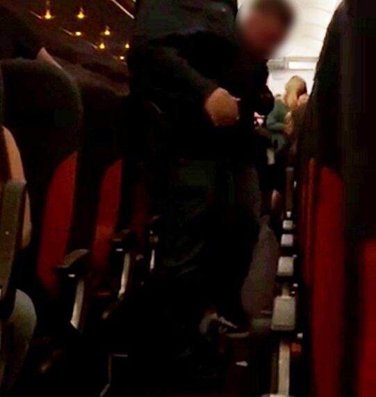 Un hombre con un uniforme oscuro escolta a un pasajero agarrando una botella de vodka por un pasillo de avión.
