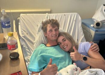 Un hombre sonriente en una cama de hospital con lesiones faciales le da un visto bueno a su novia.