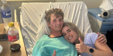 Un hombre sonriente en una cama de hospital con lesiones faciales le da un visto bueno a su novia.