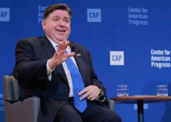 JB Pritzker, sentado, sonríe y extiende una mano mientras habla en un evento