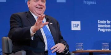 JB Pritzker, sentado, sonríe y extiende una mano mientras habla en un evento