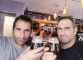 Dos hombres con cabello y rastrojo oscuros, posiblemente hermanos, sosteniendo vasos de cerveza oscura y sonriendo a la cámara en un pub.