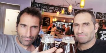 Dos hombres con cabello y rastrojo oscuros, posiblemente hermanos, sosteniendo vasos de cerveza oscura y sonriendo a la cámara en un pub.