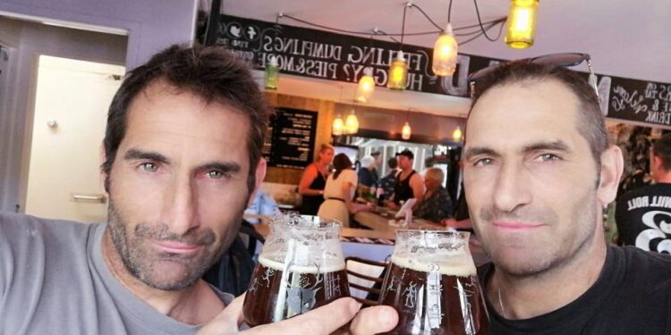 Dos hombres con cabello y rastrojo oscuros, posiblemente hermanos, sosteniendo vasos de cerveza oscura y sonriendo a la cámara en un pub.