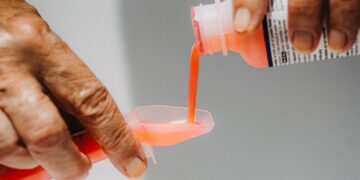 Las manos vierten medicina líquida rosa brillante de una botella en una pequeña taza de plástico.