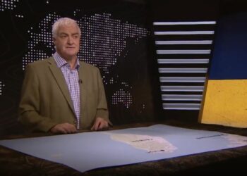 Un hombre con chaqueta de traje se para frente a un mostrador de redacción con un mapa de América del Sur y un telón de fondo que muestra un mapa mundial y una imagen agrietada de las banderas de Ucrania y Rusia.