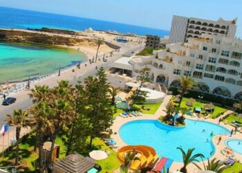 Una vista aérea de un complejo con dos piscinas, palmeras y un camino que conduce a una playa y al océano.