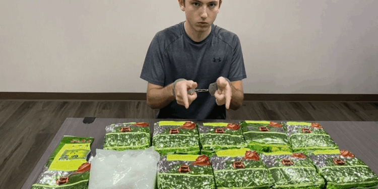Un joven con esposas se sienta detrás de una mesa con múltiples paquetes verdes y una bolsa de sustancia blanca.