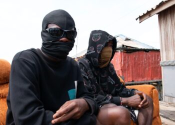 Dos personas con caras y sudaderas con capucha se sientan en un sofá naranja al aire libre.