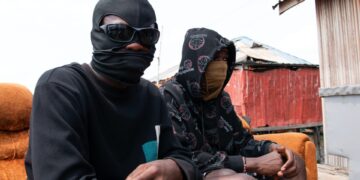 Dos personas con caras y sudaderas con capucha se sientan en un sofá naranja al aire libre.