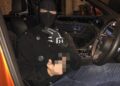 Una persona con pasamontañas negro, chaqueta negra y guantes, sentada en el asiento del conductor de un automóvil naranja, sostiene un objeto borroso en una mano.