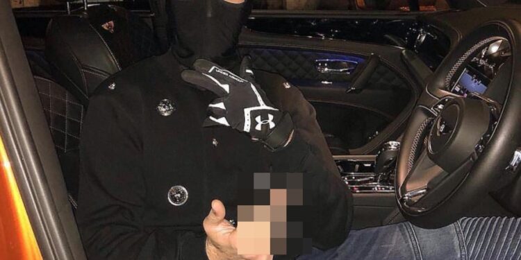 Una persona con pasamontañas negro, chaqueta negra y guantes, sentada en el asiento del conductor de un automóvil naranja, sostiene un objeto borroso en una mano.