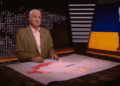 Un hombre presentando en un set de noticias con un mapa de Ucrania que muestra áreas de conflicto y una bandera rasgada de Ucrania y Rusia a la derecha.