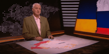Un hombre presentando en un set de noticias con un mapa de Ucrania que muestra áreas de conflicto y una bandera rasgada de Ucrania y Rusia a la derecha.