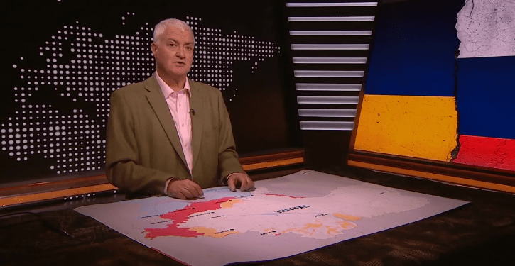 Un hombre presentando en un set de noticias con un mapa de Ucrania que muestra áreas de conflicto y una bandera rasgada de Ucrania y Rusia a la derecha.