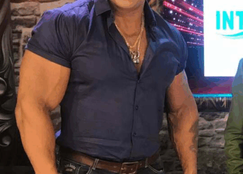 Un hombre musculoso con camisa azul, jeans y cadena de oro.