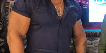 Un hombre musculoso con camisa azul, jeans y cadena de oro.