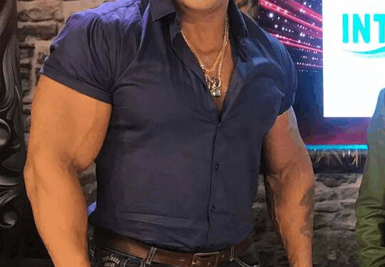 Un hombre musculoso con camisa azul, jeans y cadena de oro.
