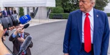 El presidente lleva un traje azul y una corbata roja y se ve caminando al lado de los medios. Los medios de comunicación están señalando cámaras al presidente.