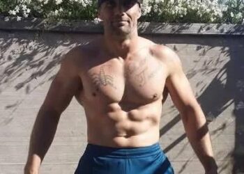 Hombre vestido con gorra de béisbol de los New York Yankees y pantalón corto azul, mostrando su torso con tatuajes.