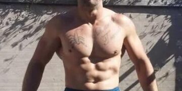 Hombre vestido con gorra de béisbol de los New York Yankees y pantalón corto azul, mostrando su torso con tatuajes.