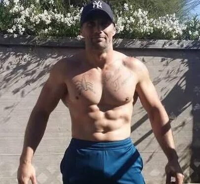 Hombre vestido con gorra de béisbol de los New York Yankees y pantalón corto azul, mostrando su torso con tatuajes.