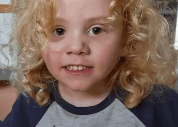 Un niño pequeño con cabello rubio rizado sonríe ligeramente a la cámara, con una camisa gris con el texto "Mi mamá" y un personaje de cerdo rosa.