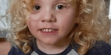 Un niño pequeño con cabello rubio rizado sonríe ligeramente a la cámara, con una camisa gris con el texto "Mi mamá" y un personaje de cerdo rosa.