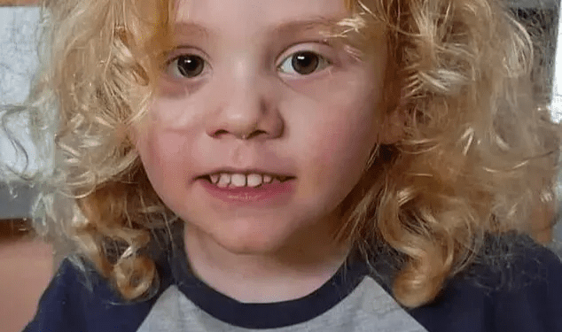 Un niño pequeño con cabello rubio rizado sonríe ligeramente a la cámara, con una camisa gris con el texto "Mi mamá" y un personaje de cerdo rosa.