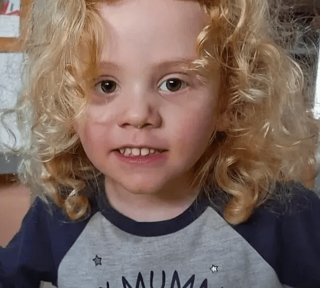 Un niño pequeño con cabello rubio rizado sonríe ligeramente a la cámara, con una camisa gris con el texto "Mi mamá" y un personaje de cerdo rosa.