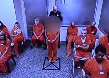 Una mujer con un mono naranja mira hacia adelante mientras un guardia de seguridad está detrás de ella en una habitación con otras personas con monos naranjas.