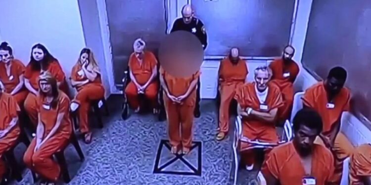 Una mujer con un mono naranja mira hacia adelante mientras un guardia de seguridad está detrás de ella en una habitación con otras personas con monos naranjas.