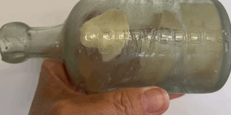 Una mano sosteniendo una botella de vidrio transparente con la palabra "CL Wedel's" grabado en relieve, con papel enrollado de color claro visible en el interior.