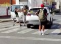 Un hombre con una camisa negra y pantalones cortos blancos argumenta con otro hombre con una camisa y un sombrero marrones, que está parado junto a un auto blanco en un cruce de cebra.