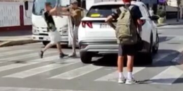 Un hombre con una camisa negra y pantalones cortos blancos argumenta con otro hombre con una camisa y un sombrero marrones, que está parado junto a un auto blanco en un cruce de cebra.