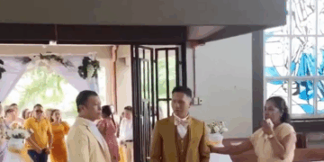 Un hombre con un traje dorado, de pie en un pasillo rojo, mira hacia una mujer con un vestido largo beige que habla por un micrófono. Otro hombre con un traje claro está a la izquierda del hombre del traje dorado, mirando hacia una procesión nupcial que entra por las puertas abiertas.