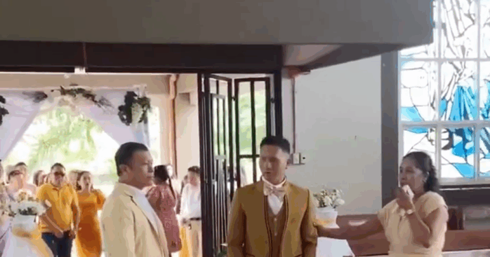 Un hombre con un traje dorado, de pie en un pasillo rojo, mira hacia una mujer con un vestido largo beige que habla por un micrófono. Otro hombre con un traje claro está a la izquierda del hombre del traje dorado, mirando hacia una procesión nupcial que entra por las puertas abiertas.