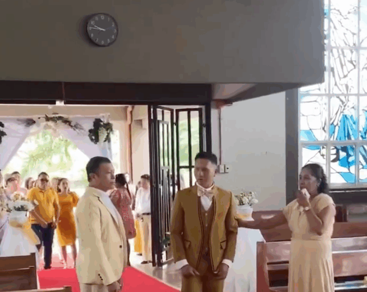 Un hombre con un traje dorado, de pie en un pasillo rojo, mira hacia una mujer con un vestido largo beige que habla por un micrófono. Otro hombre con un traje claro está a la izquierda del hombre del traje dorado, mirando hacia una procesión nupcial que entra por las puertas abiertas.