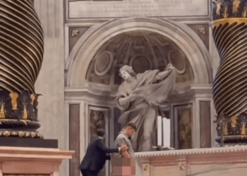 Un hombre vestido con un traje gris claro y los pantalones bajados es inmovilizado por otro hombre con un traje oscuro frente a un altar, con sus partes íntimas pixeladas.