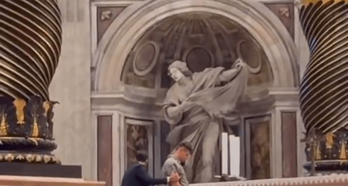 Un hombre vestido con un traje gris claro y los pantalones bajados es inmovilizado por otro hombre con un traje oscuro frente a un altar, con sus partes íntimas pixeladas.