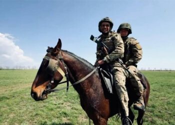 Dos soldados en camuflaje y cascos montan un caballo marrón a través de un campo cubierto de hierba.