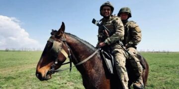 Dos soldados en camuflaje y cascos montan un caballo marrón a través de un campo cubierto de hierba.