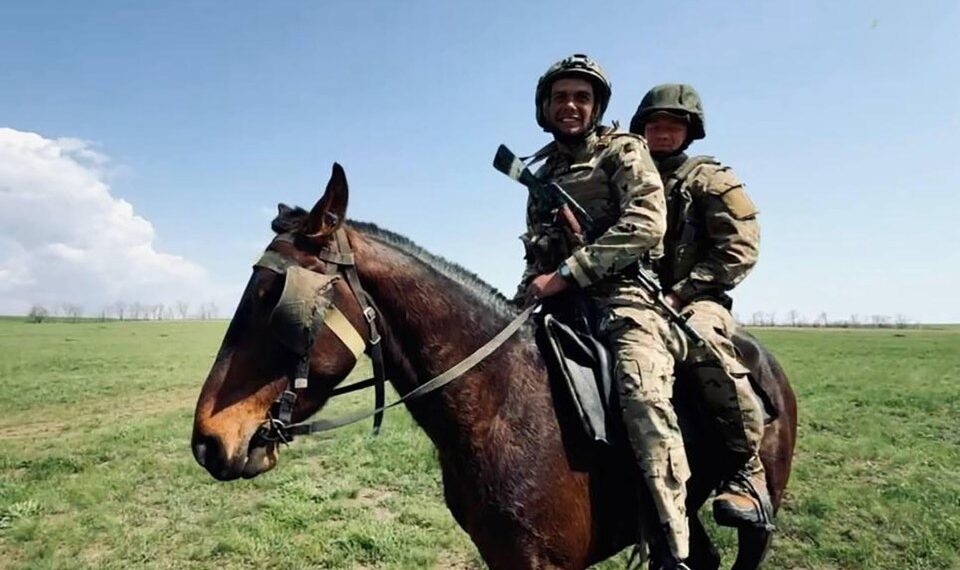 Dos soldados en camuflaje y cascos montan un caballo marrón a través de un campo cubierto de hierba.