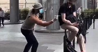 Una persona con un sombrero, cara facial y guantes que fingen inyectar a una persona sentada en un banco con una jeringa.