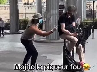 Una persona con un sombrero, cara facial y guantes que fingen inyectar a una persona sentada en un banco con una jeringa.